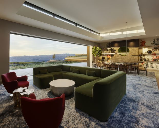 A spacious contemporary open-plan living and dining area with expansive picture-window glazing overlooking a mountainous landscape. The room features a large deep-green sectional sofa, red lounge chairs, a round coffee table, patterned blue-grey rug, sculptural pendant lighting, and a dining area adjoining a kitchen with dark cabinetry, open shelving, and a stone-look backsplash. Clerestory glazing and a long linear roof skylight bring in abundant natural light.