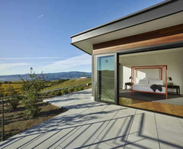 Contemporary single-level pavilion with a flat roof and broad retractable glazed opening, viewed from a generous concrete terrace. The open bedroom has a minimalist four-poster bed and dark flooring, while the exterior overlooks landscaped grounds and a distant mountain valley under clear blue skies.