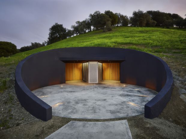 A partially subterranean, circular concrete-and-dark-render structure set into a grassy hillside. The recessed curved forecourt has a smooth concrete floor with integrated ground lights and a central timber-clad entrance, flanked by timber doors and a perforated metal panel. Trees and a sloping lawn rise behind the structure under an overcast sky.