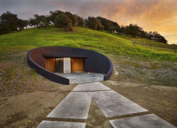 A partially earth-sheltered, circular dark-clad structure embedded into a grassy hillside. A concrete path leads to a recessed entrance with warm timber doors and a central pale metal door, set within a curved dark wall and concrete forecourt. The surrounding rural landscape includes rolling grass, mature trees, fencing, and dramatic sunset light.