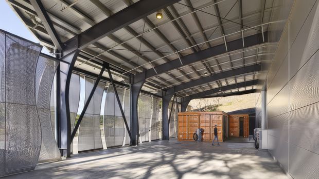 A large covered industrial-style outdoor passage or loading area with exposed dark steel beams, corrugated metal roofing, perforated metal screening along the left side, and a ribbed metal-clad wall on the right. Timber storage or service structures sit at the far end, with strong filtered daylight and tree shadows across the concrete floor.