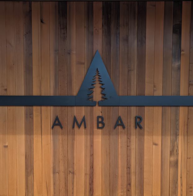 Close-up of a rustic timber-clad wall featuring a dark horizontal metal band, a stylised mountain-and-evergreen emblem, and raised lettering spelling “AMBAR.” The warm vertical wood boards and dark graphic detailing create a natural, lodge-inspired branded facade or feature wall.