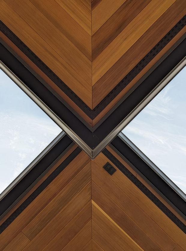 Close-up upward-looking architectural detail of a sharp building corner or folded roofline. Warm timber panels are arranged in diagonal and chevron-like formations, bordered by dark metal bands and narrow perforated ventilation strips. Reflective metal trim and pale sky are visible around the geometric junction, creating a highly graphic, symmetrical composition.