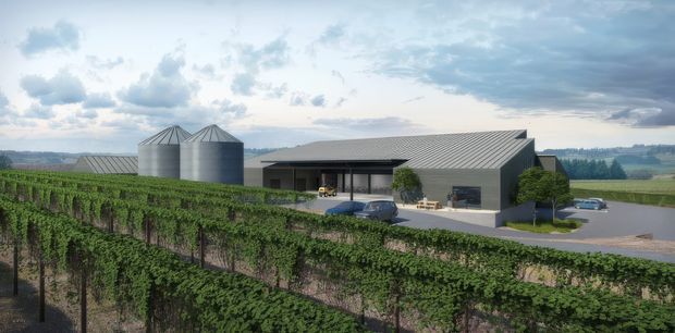 Wide exterior view of a contemporary agricultural or winery facility set among orderly vineyard rows. The building has a simple low-profile form with dark grey metal-clad walls, a large pitched metal roof, deep covered entry or loading area, and extensive paved forecourt. Several cylindrical grain or storage silos sit beside the building, with rolling rural farmland visible beyond.