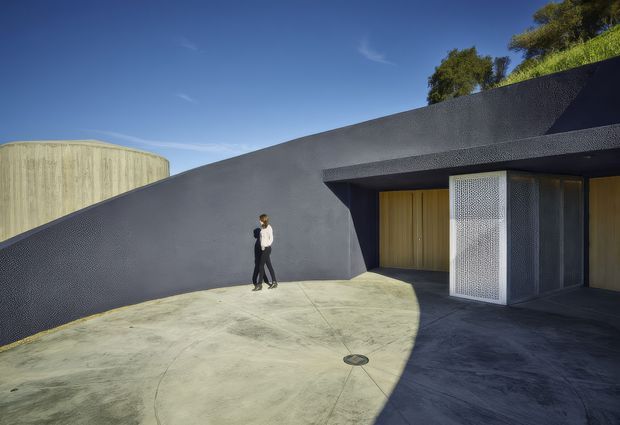 A sculptural, low-profile dark grey building exterior with a sharply angled wall and broad flat roof canopy. The entrance features warm timber doors and a perforated metal screen enclosure, opening onto a curved pale concrete forecourt. A large cylindrical concrete form is visible behind the sloping wall, with planted greenery above the structure. The image has a strong geometric, minimalist architectural character under clear daylight.