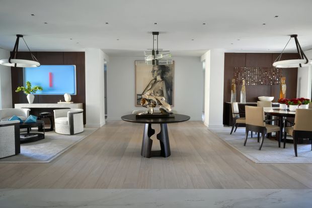 A refined open-plan interior combining a living room and dining area. The space features pale timber flooring, white walls and ceiling, dark timber feature panels, contemporary pendant lighting, a sculptural central console table, a wall-mounted television, modern seating, and a formal dining setting. A large artwork and decorative objects add visual interest.
