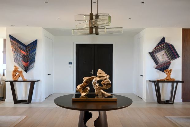 A bright, refined residential entry hall with a symmetrical composition. Black double front doors are centered on a white wall, flanked by narrow doorways and console tables displaying sculptural artwork. A round dark table with a polished gold abstract sculpture anchors the foreground, while a geometric tiered glass-and-metal pendant hangs above. Light timber flooring, white walls, dark accents, and colourful geometric wall art create a contemporary gallery-like atmosphere.