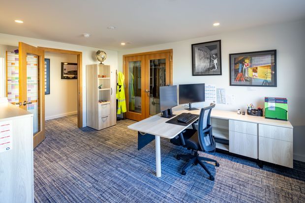 Bright modern office interior with two computer workstations, ergonomic office chairs, white desks, low storage cabinetry, framed artwork, shelving, and timber-framed glazed doors. The room has a neutral palette, recessed ceiling lights, and a blue-grey patterned carpet, with an adjoining corridor and open doorway visible.