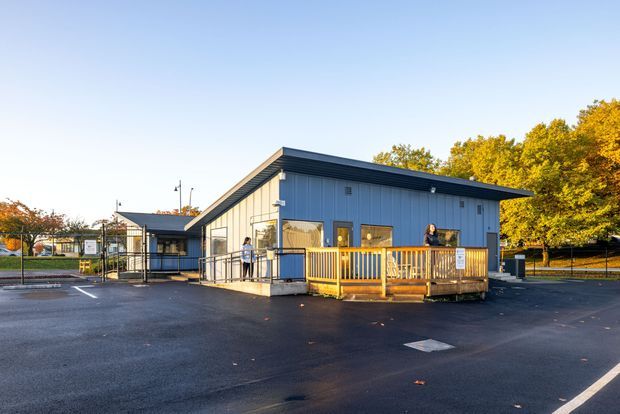 Exterior view of a compact single-storey commercial or community building with a simple sloped metal roof, blue-grey vertical cladding, windows, entry ramp, and a small timber-railed deck. The building sits beside a paved parking area and fenced grounds, with autumn trees in the background.