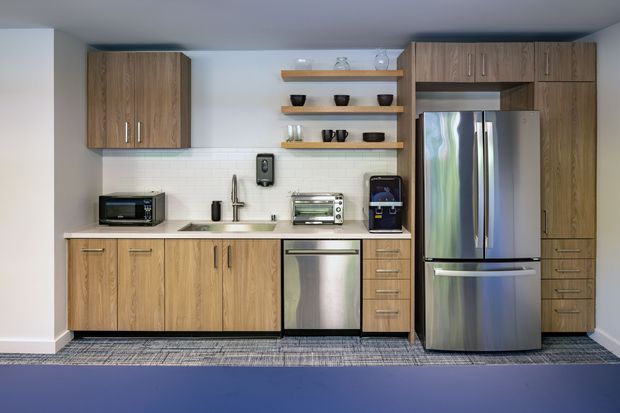 Compact contemporary kitchenette with full-height wood-grain cabinetry, pale stone-look benchtop, white tiled splashback, stainless-steel appliances, open timber shelves, and integrated sink. The linear layout includes an under-bench dishwasher, oven, refrigerator, microwave, toaster oven, coffee machine, and drawers. Neutral finishes and clean lines create a practical, modern workspace.