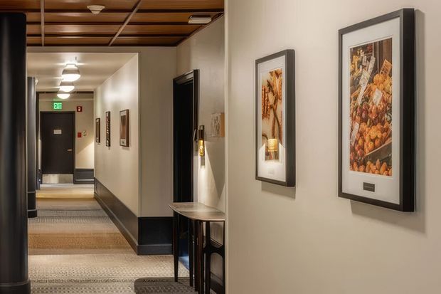 A warmly lit interior corridor or hotel-style hallway with framed artwork along cream walls, patterned carpet flooring, dark trim and doors, and a distinctive timber slatted ceiling with recessed lighting. A narrow console table and wall-mounted fixtures line the corridor, which extends toward an illuminated exit.