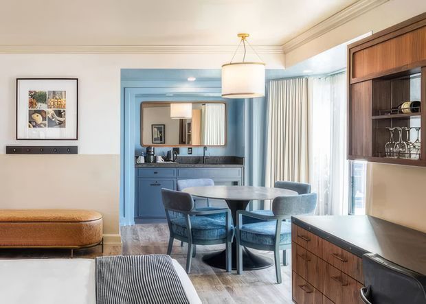 A refined open-plan dining and living area with a compact blue kitchen in the background. The room features a round dining table with upholstered chairs, a large drum pendant light, built-in dark timber cabinetry with shelving and a dark benchtop, blue painted kitchen cabinetry, striped upholstered seating, framed artwork, and full-height curtains. The space has a warm, sophisticated mid-century-inspired character with strong blue, cream, timber, and grey tones.
