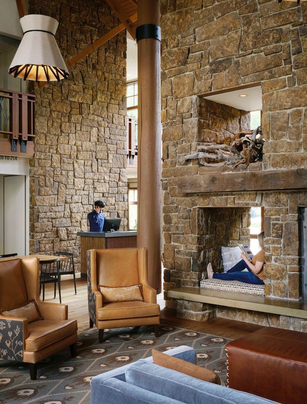 A rustic, lodge-like double-height living room dominated by exposed rough stone walls and a massive stone fireplace with built-in seating. Leather armchairs and sofas, a patterned rug, timber structural columns and beams, and a large woven pendant create a warm, rugged interior. A small reception or kitchen counter is visible in the background, with a person seated beside the fireplace.