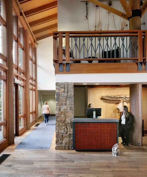 A spacious double-height reception lobby with a prominent timber post-and-beam structure, extensive floor-to-ceiling timber-framed glazing, exposed pitched roof framing, and a mezzanine level with decorative dark metal balustrading. The reception desk has a dark stone-like counter and patterned timber front, while a rugged natural-stone column, timber-clad wall, and cylindrical timber post add texture. A blue-grey carpeted circulation zone contrasts with broad timber flooring. The space has a warm, lodge-inspired architectural character and serves as a reception or commercial entry area.