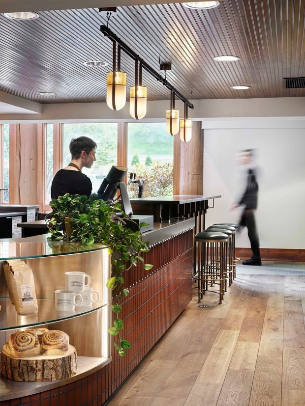 Interior photograph of a stylish café or hospitality counter, featuring a long terracotta-toned tiled bar with dark countertop, brass-toned curved display shelving, coffee equipment, potted greenery, and tall bar stools. Warm timber flooring and wall framing contrast with a ribbed metallic ceiling and a linear track of sculptural pendant lights. Large windows bring in natural light and views of greenery, while a blurred person moves through the space.