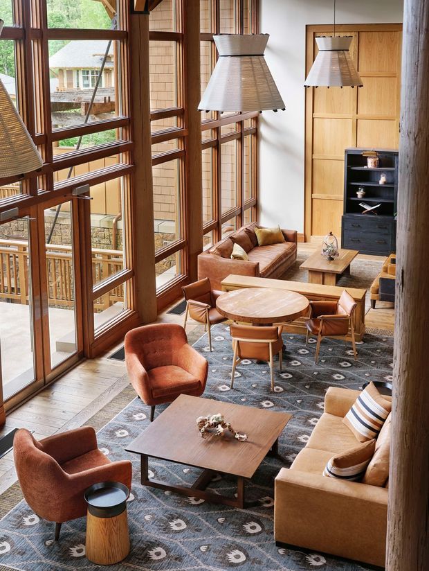 A spacious, double-height living room with a loft-like upper viewpoint, extensive floor-to-ceiling timber-framed glazing, exposed heavy timber posts, and warm natural finishes. The room contains leather and upholstered seating arranged around a round dining or occasional table and a square coffee table, with a timber built-in door and dark shelving at the rear. Large woven pendant lights hang over the seating area, and a patterned grey rug anchors the furniture. The glazing looks onto a wooded, multi-level exterior setting.