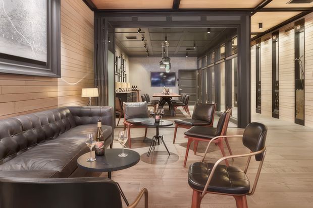 A sophisticated open-plan lounge and dining area with a strong mid-century influence. The room features a tufted dark leather sofa, leather lounge chairs with timber and metal frames, round black coffee tables, a dining table, and a television at the rear. Warm timber wall panelling and timber flooring are paired with dark-framed glazed partitions and doors. Track lighting and pendant fixtures provide illumination, while a corridor or glazed room extends along the right side.