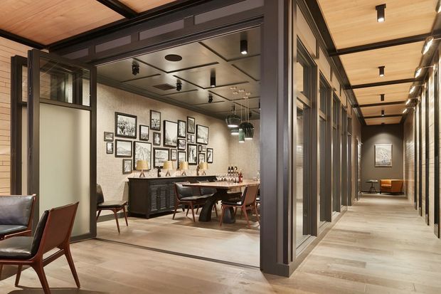 A refined interior showing a dining and lounge area opening onto a long hallway. The space features light timber flooring and ceiling boards, dark charcoal structural framing and trim, extensive glazed partitions and doors, a long dark sideboard, a dining table with leather dining chairs, framed artwork arranged gallery-style, and warm integrated and track lighting. A seating area is visible at the far end of the corridor.
