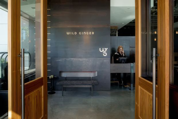 Entrance view into the Wild Ginger restaurant, framed by open timber-and-glass doors. A dark metal feature wall with gold lettering and logo sits behind a compact upholstered bench, while a reception counter and staff member are visible deeper inside. The space has polished concrete flooring, dark finishes, and warm timber detailing, creating a refined contemporary hospitality interior.