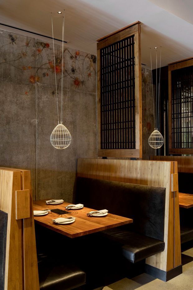 A warmly lit restaurant dining area with built-in booth seating, richly grained timber partitions and tables, black leather upholstery, and decorative pendant lights. The walls feature textured concrete-like panels with an abstract floral motif, while dark vertical screens and mirrors create privacy and depth. The space has a refined, atmospheric contemporary interior with strong Japanese-inspired detailing.