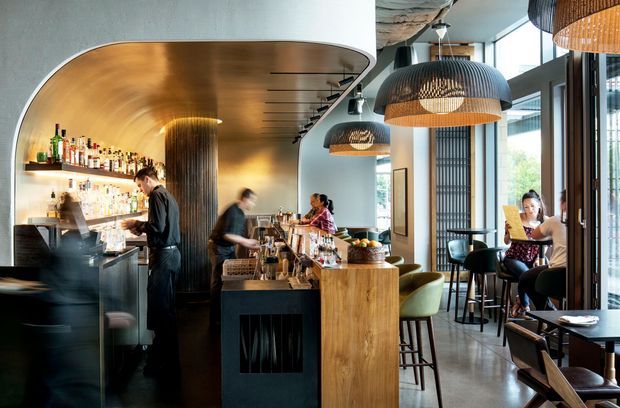 A refined contemporary bar and dining space with a curved metallic feature ceiling and back bar, dark stone or metal bar counters, timber-clad counter fronts, and large pendant lights with woven shades. Bartenders work behind the central bar while patrons sit at small tables and high stools beside full-height windows. The space has a sophisticated hospitality atmosphere with warm lighting and a mix of dark, timber, and green upholstered furnishings.