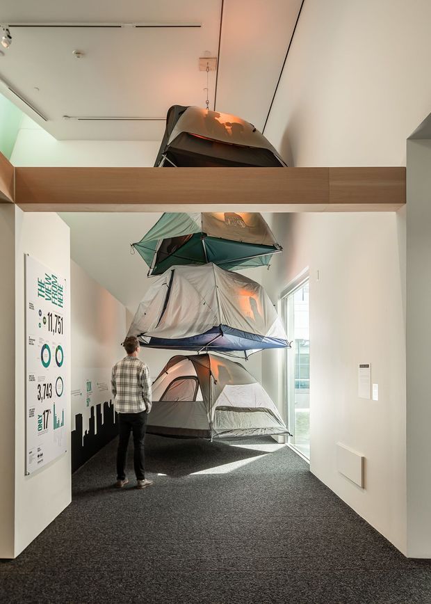 A narrow, contemporary interior passage or gallery with white walls, dark carpet flooring, and a suspended timber-clad horizontal beam. Several camping tents are dramatically stacked and suspended through the space as an exhibition or installation, with an informational graphic panel on the left wall and daylight entering through glazing on the right.