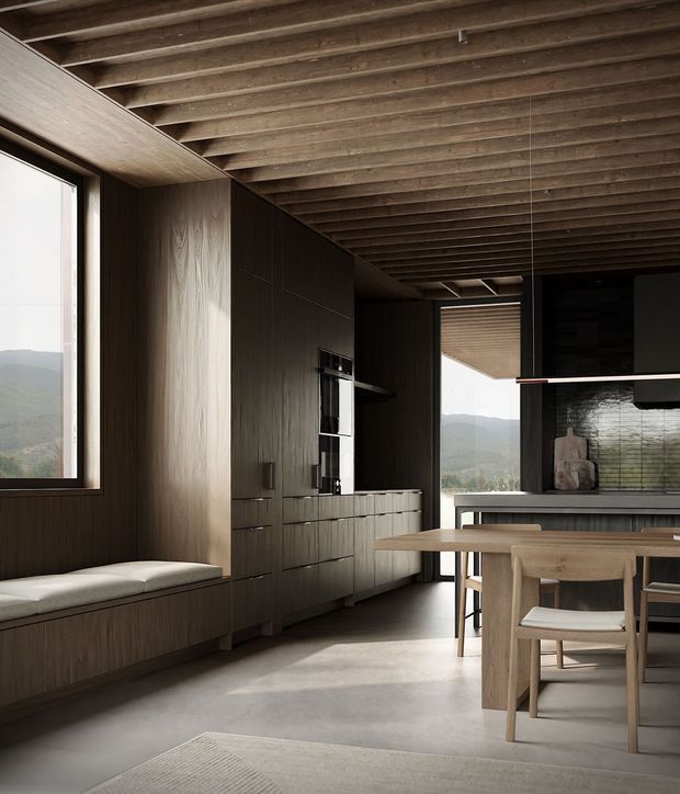 A refined open-plan kitchen and dining area with dark timber cabinetry, a built-in window seat, concrete flooring, and a timber dining table. Exposed timber ceiling joists create a strong linear architectural feature, while large windows and an open glazed doorway frame a mountainous landscape. The kitchen includes a dark island or counter, integrated appliances, and a textured dark backsplash.