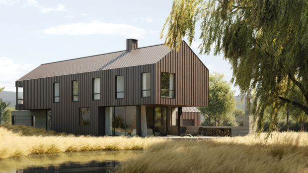 A contemporary two-storey house set in a landscaped rural or suburban setting. The building has a dark vertically ribbed metal-clad facade, a pitched gable roof, tall narrow windows, and a partially covered ground-level outdoor living area. Ornamental grasses and mature weeping trees surround the home, with distant hills visible beyond.