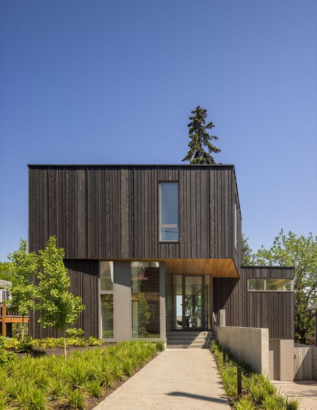 Exterior photograph of a contemporary multi-level home with a strong rectangular form, dark vertical timber cladding, expansive glazing, and a projecting upper volume over the recessed entry. A concrete walkway and low retaining wall lead through landscaped planting to the entrance. The home has a flat roof and a restrained, modern material palette.