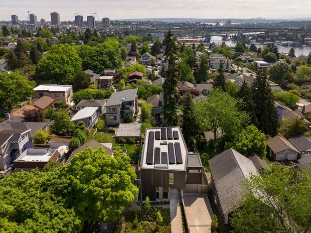 Aerial view over a leafy suburban neighbourhood, with a modern multi-storey house featuring a flat roof and multiple rooftop solar panels in the foreground. Dense tree cover surrounds a mix of detached homes, while a broad body of water, bridges or waterfront infrastructure, and a high-rise urban skyline are visible in the distance.
