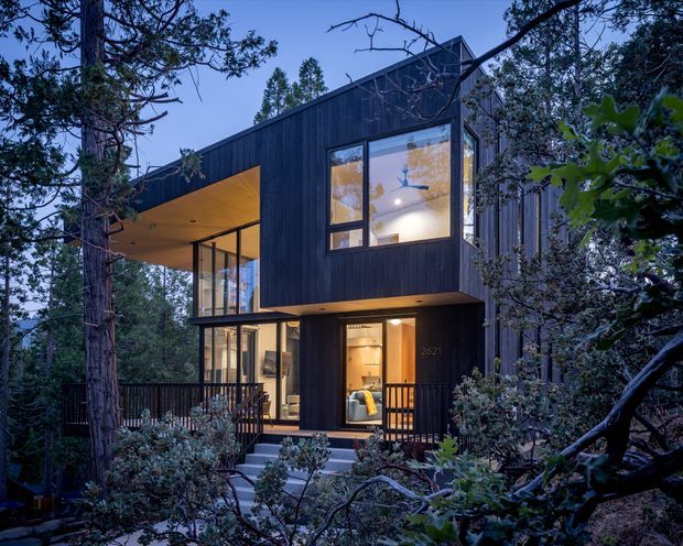 Exterior view of a contemporary two-storey home set among mature trees in a wooded setting. The house has dark vertical cladding, a sharply folded asymmetrical roofline with a deep cantilevered upper level, extensive floor-to-ceiling glazing, and a warm timber-lined recessed entry and balcony soffit. Exterior steps and decking lead toward the illuminated entrance.