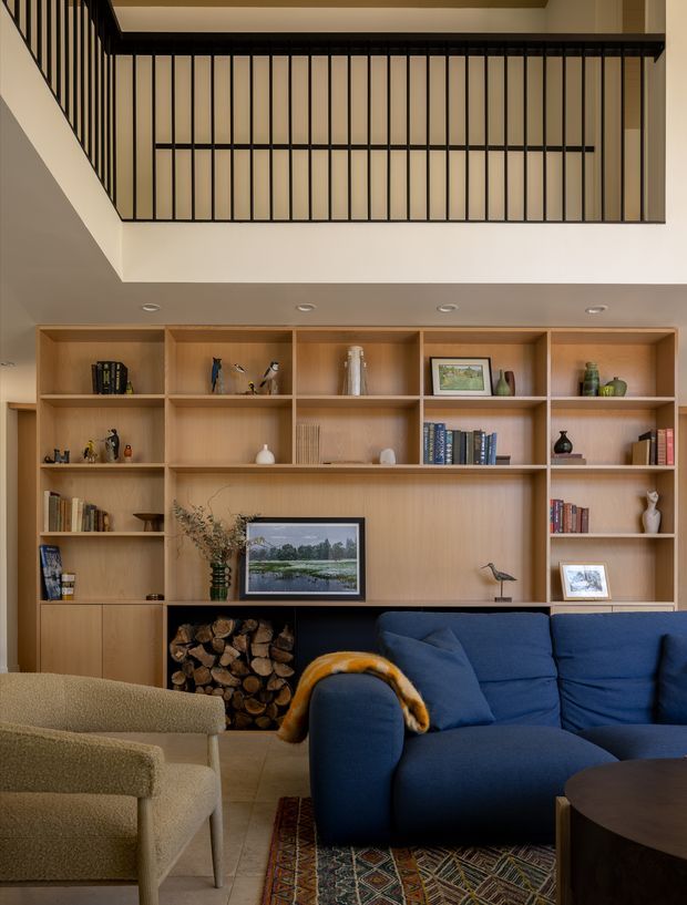 A warm, refined living room with full-height built-in timber shelving and cabinetry, a dark fireplace or log-storage niche, and a prominent navy-blue sofa. An upper-level void is enclosed by a black vertical metal balustrade. Books, ceramics, framed artwork, and sculptural objects fill the shelving, while a patterned rug and neutral armchair complete the comfortable interior.