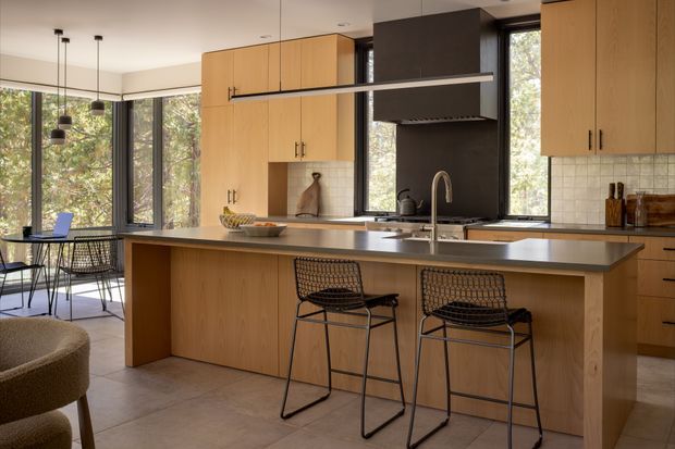 A warm, contemporary open-plan kitchen with extensive light timber cabinetry, a large island with a dark stone benchtop, integrated cooktop and sink, and two black wire bar stools. A dark feature rangehood wall contrasts with the white square-tile splashback. Floor-to-ceiling corner glazing opens the kitchen and adjacent dining area to leafy views, while pendant lights hang above the dining table.