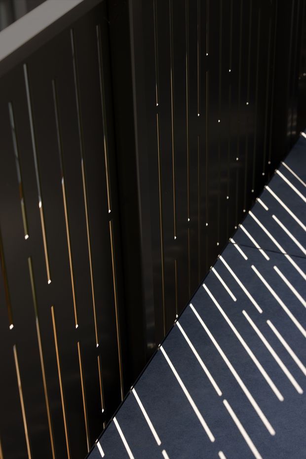 Abstract architectural detail of a dark, glossy vertical screen or facade with thin linear metallic accents and small points of light. Strong sunlight creates repeated bright diagonal shadow stripes across a dark paved or tiled surface, producing a dramatic, minimalist geometric composition.