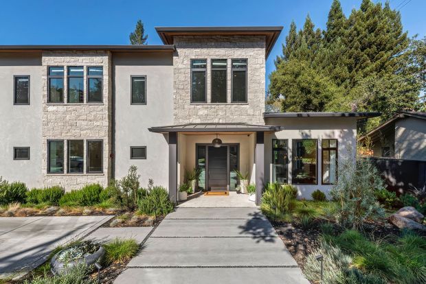 Front exterior view of a contemporary multi-level residence with a flat roof, pale stone masonry and smooth rendered walls. Dark-framed windows, a recessed entry porch, broad concrete pathway, and landscaped drought-tolerant planting define the facade.