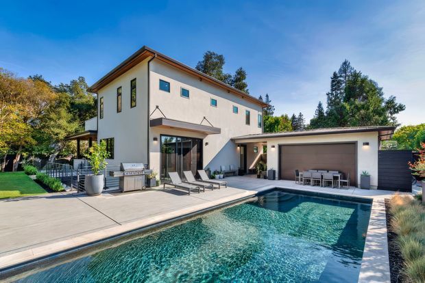 Exterior view of a contemporary multi-level residence with light rendered walls, dark trim, and a low-pitched roof. The home opens onto a broad paved patio and rectangular swimming pool, with an attached single-storey garage and landscaped planting around the property.
