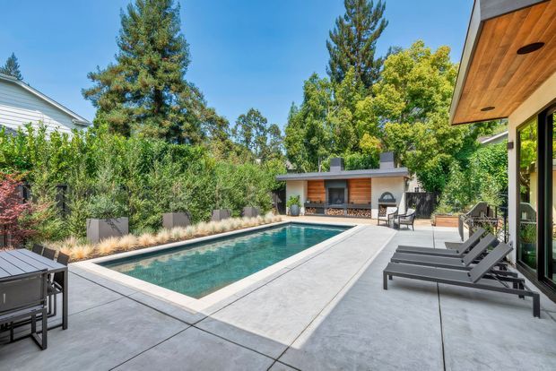 Contemporary landscaped backyard with a rectangular swimming pool surrounded by a broad concrete terrace. The space includes black-framed glazing, timber-lined soffits, modern sun loungers, outdoor dining furniture, and a compact poolside pavilion with timber cladding, a built-in barbecue or outdoor kitchen, and seating. Mature planting creates a private, leafy setting.