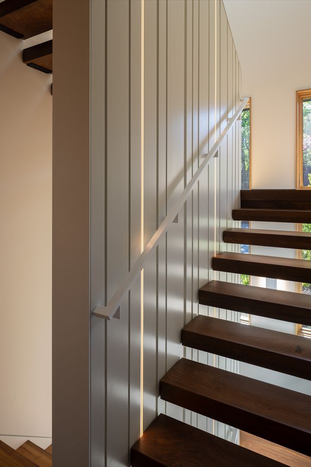Interior view of a modern open-riser staircase with dark timber treads cantilevered beside a tall vertically panelled wall. A slim integrated linear light runs up the wall, while a simple pale handrail follows the stair line. Timber ceiling beams and windows with timber frames are visible, creating a warm contemporary stairwell with strong vertical and horizontal lines.