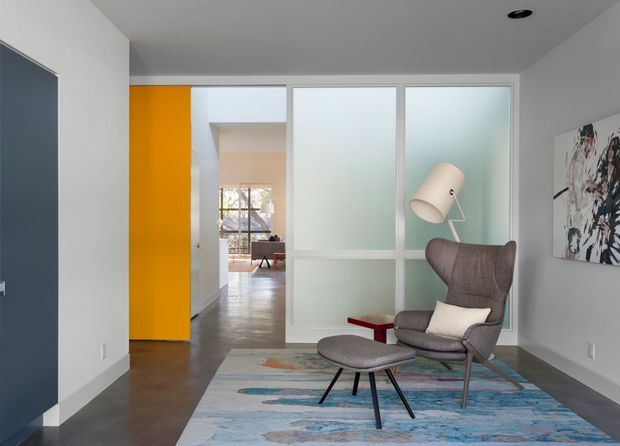 A contemporary sitting area with a sculptural upholstered lounge chair and matching ottoman, positioned on a large abstract blue-toned rug. A tall floor lamp and small red side table create accents beside the seating. The room has pale grey walls, dark polished flooring, a vivid orange door, and a frosted-glass partition separating it from a brighter adjoining space.