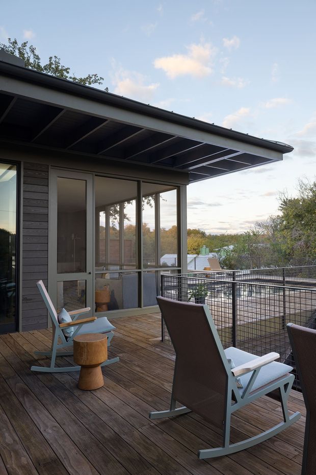 A contemporary elevated timber deck or terrace beside a dark-clad house, furnished with modern rocking lounge chairs and a small wooden side table. The covered roof overhang has exposed dark structural framing, while full-height glazed doors and windows connect the terrace to the interior. A wire-mesh balustrade overlooks leafy surroundings.