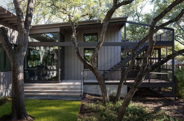 Exterior view of a contemporary multi-level residence integrated among mature trees. The dark vertical-clad facade has extensive floor-to-ceiling glazing, a low-pitched roof, and an elevated external stair and terrace with slim dark metal framing and mesh balustrades. Concrete entry steps and landscaped garden beds occupy the foreground.