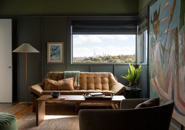 A moody, mid-century-inspired living room with dark green-grey paneled walls, a large framed artwork, and a broad window overlooking a leafy urban landscape. The room contains a tufted caramel leather sofa, dark upholstered lounge chair, wooden rectangular coffee table, slim floor lamp, patterned rug, indoor plant, and a large colorful wall artwork. Natural daylight from the window balances the dark interior palette.