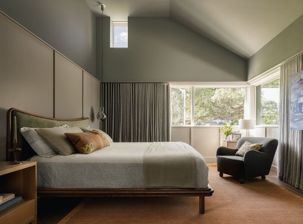 A calm, refined bedroom with a high sloping ceiling and clerestory window. The room features a low timber bed with upholstered headboard, built-in floor-to-ceiling cabinetry, warm wood flooring, neutral green-grey walls, full-height curtains, and a large corner window overlooking trees. A dark upholstered armchair and small side table occupy the sitting area.