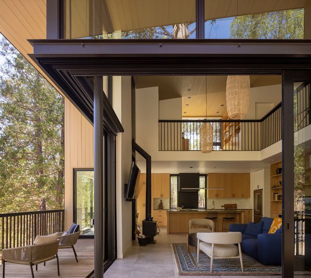 A tall, open-plan contemporary living area viewed through a wide glazed opening to a covered timber deck. The interior features a double-height ceiling, mezzanine walkway with black vertical balustrading, extensive floor-to-ceiling glazing, pale timber cabinetry, a compact kitchen, stone-look flooring, and a blue sectional sofa. Warm timber surfaces and woven pendant lights contrast with dark metal window frames and railings.