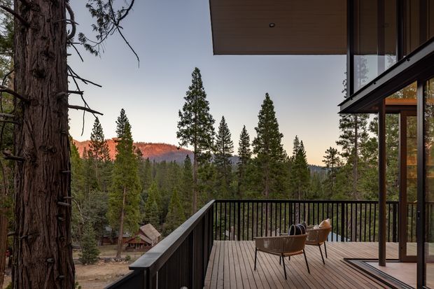A contemporary timber-clad deck or balcony viewed from a sheltered outdoor living area, overlooking a dense conifer forest and distant wooded hills. The deck has dark metal or painted balustrades, timber decking, glazed sliding doors, and a broad timber-lined roof soffit. Outdoor lounge chairs and a small table create a quiet, elevated retreat integrated with the surrounding natural landscape.