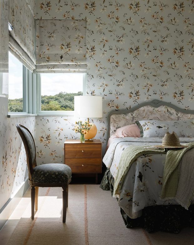 A softly lit bedroom with floral wallpaper covering the walls and ceiling, a large window with a Roman shade, and a vintage-style upholstered bed with floral bedding. A timber bedside chest, table lamp, upholstered wooden chair, vase of flowers, and layered green throw create a warm, traditional and cozy atmosphere. The room overlooks leafy landscape through the window.