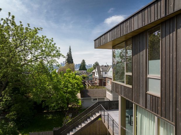 Exterior architectural view of a contemporary multi-level home clad in dark vertical timber boards, with extensive floor-to-ceiling glazing, projecting roof eaves, and elevated timber decks and stairs. The house sits in a leafy suburban setting with neighbouring homes and a distant urban skyline visible beyond.