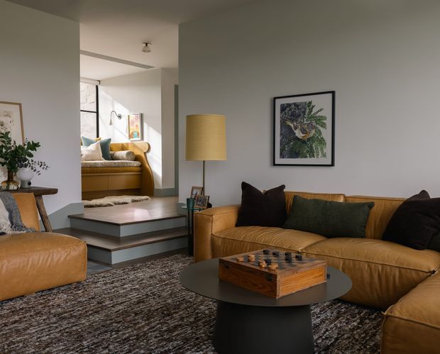 Warm, layered living room with a large cognac leather sectional sofa, matching leather armchair, dark round coffee table, patterned area rug, framed artwork, and a brass-toned floor lamp. The room opens through a short set of steps to a brighter adjoining sitting area with additional leather seating and artwork. Neutral plaster walls and simple ceiling detailing create a relaxed, refined interior.
