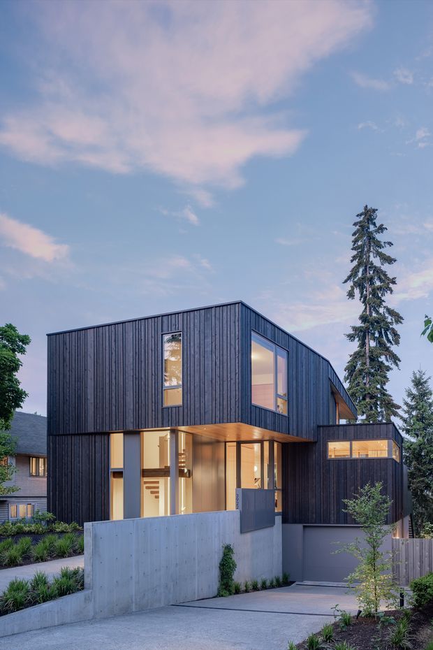 Exterior view of a contemporary multi-level residence with a strongly geometric, angular form. The facade is clad in dark vertical timber boards, with large glazed openings, warm interior lighting, projecting upper volumes, and exposed concrete boundary walls. A concrete driveway and landscaped front garden lead to the entrance and garage. Mature trees and a neighbouring house are visible around the property.