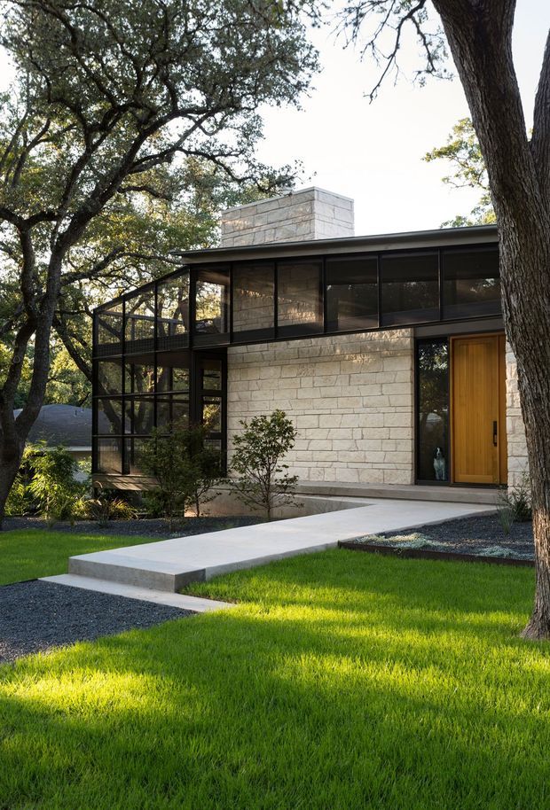 Exterior view of a low-profile modern house set within a mature, heavily planted landscape. The facade combines pale coursed masonry with dark slender metal-framed glazing, including a screened or enclosed upper-level terrace at left. A tall projecting masonry chimney rises above the flat roofline, while a warm timber front door opens onto a broad concrete walkway through a neatly manicured lawn and gravel garden beds.